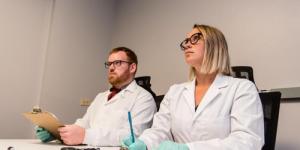 Two people sitting in lab coats with clip pads and pens observing something offscreen